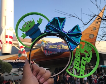 May include: Green and blue 3D printed Minnie Mouse ears with a green alien character from Toy Story on one ear and the words "Pizza Planet" on the other ear. The ears are being held by a person in front of a rocket ship.