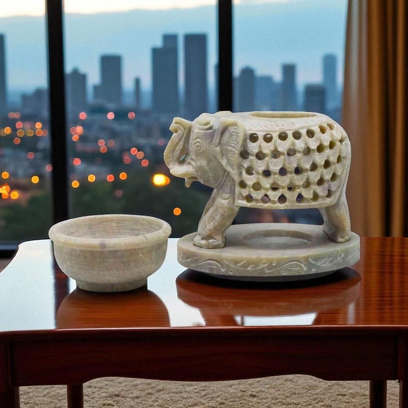 May include: A carved stone elephant sculpture and a bowl. The elephant has a hollowed body with a circular opening. The bowl is round and sits beside the elephant. The objects are light beige in colour.