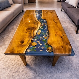 Pode incluir: Mesa de centro retangular de madeira com um design de rio de resina azul. O rio é preenchido com conchas, estrelas do mar e outros elementos marinhos. A mesa tem um acabamento natural de grão de madeira e pernas de madeira anguladas.