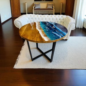 Op de afbeelding: Ronde houten salontafel met een uniek epoxyharsontwerp met oceaanthema. De tafel heeft een natuurlijke houtnerf afwerking aan de ene helft en een blauw en wit harsontwerp dat op golven lijkt aan de andere kant. De tafel wordt ondersteund door een zwart metalen onderstel.