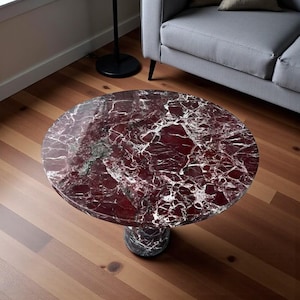 Puede incluir: Una mesa de centro redonda con una tapa de mármol rojo con vetas blancas. La mesa tiene una base cilíndrica única hecha de mármol similar. La mesa está sobre un suelo de madera.