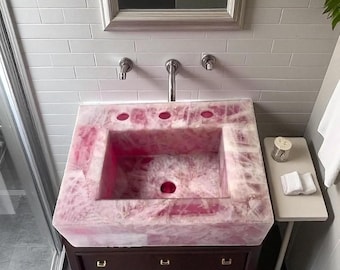 Puede incluir: Un lavabo de baño de cuarzo rosa con una cubeta rectangular y tres orificios para el grifo. El lavabo está colocado sobre un mueble de madera marrón oscuro con tres cajones. El fondo incluye una pared de azulejos blancos y un grifo plateado.