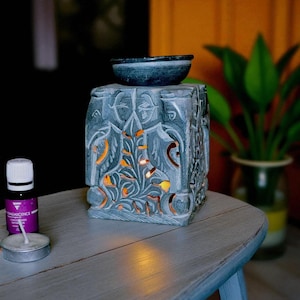 Puede incluir: Un difusor de aceites esenciales de piedra gris con un diseño floral tallado. El difusor tiene un pequeño cuenco en la parte superior y patrones recortados que permiten que la luz brille. Una pequeña botella de aceite esencial y una vela de té están al lado.