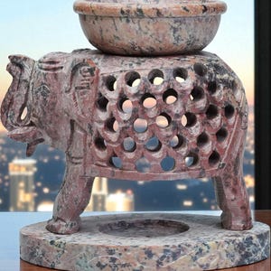 May include: A carved stone elephant oil burner with a bowl on top. The elephant has a detailed design with cut-out circles. The base is a round, speckled stone. The color is a mix of red, brown, and grey.