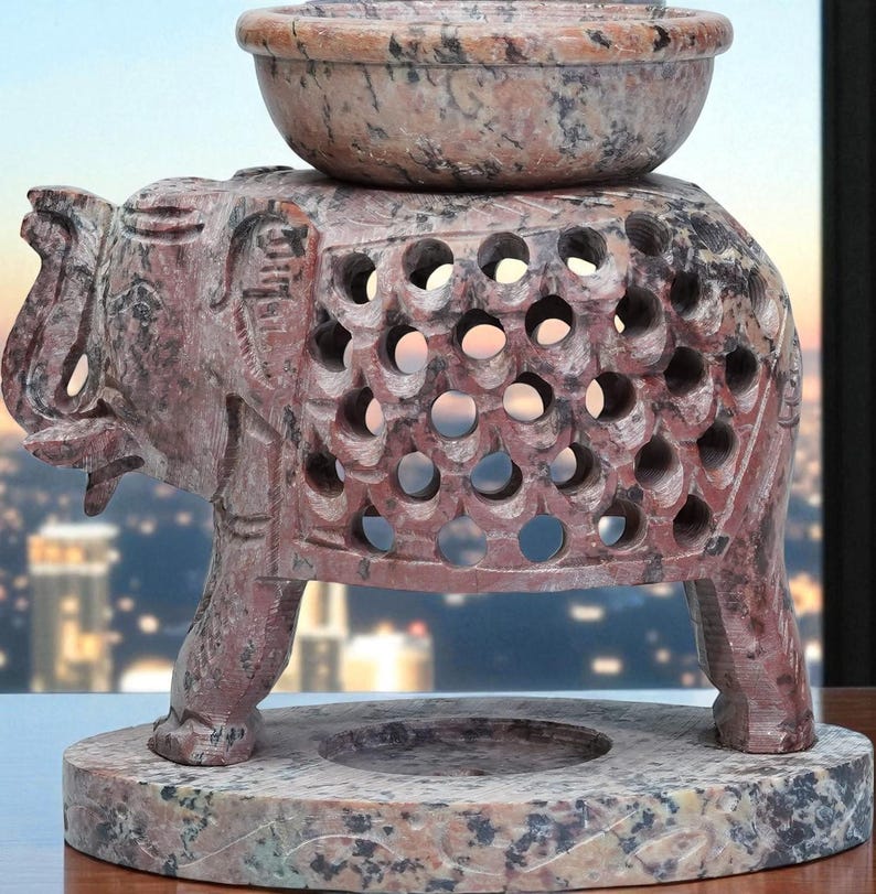 May include: A carved stone elephant oil burner with a bowl on top. The elephant has a detailed design with cut-out circles. The base is a round, speckled stone. The color is a mix of red, brown, and grey.