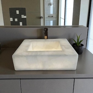 May include: A modern, square, white marble bathroom sink with a built-in faucet. The sink is illuminated from within, creating a soft glow. A small potted plant sits to the side, adding a touch of greenery to the bathroom decor.