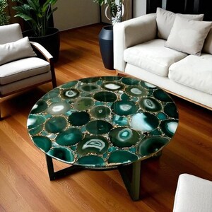 Puede incluir: Mesa de centro redonda con tapa de piedra de ágata verde. La mesa presenta un mosaico de piedras de ágata verdes y blancas con detalles dorados. La mesa tiene una base verde oscuro y está sobre un suelo de madera.