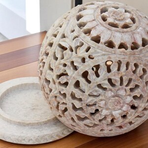 May include: A carved stone sphere with intricate floral patterns and a matching circular base. The sphere is off-white with a detailed openwork design. The base is a flat, round dish. The decorative object is suitable for home decor.