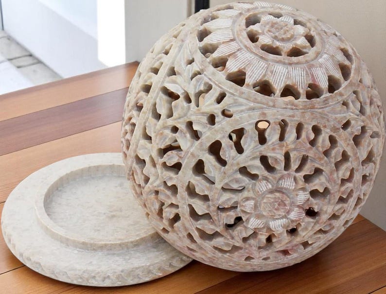 May include: A carved stone sphere with intricate floral patterns and a matching circular base. The sphere is off-white with a detailed openwork design. The base is a flat, round dish. The decorative object is suitable for home decor.