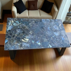 May include: A rectangular table with a dark blue, mosaic-like surface. The table has black legs and is surrounded by two chairs with brown seats and backs. The table is in a room with a sofa and a window.