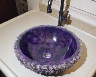 May include: A round, purple amethyst stone sink with a rough, textured edge. The sink is set in a white countertop with a dark metal faucet. The sink's interior has a polished, glossy finish.
