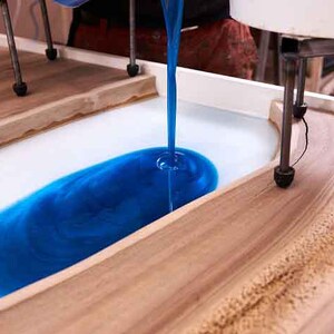 May include: A woodworking project in progress, showing blue resin being poured into a wooden mold. The mold has a natural wood grain and a white base. The resin is being poured from a machine, creating a striking contrast.