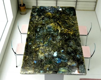 May include: A rectangular dining table with a polished, multi-colored stone top. The stone features shades of green, blue, and gold. The table is supported by a light gray base. Two pink chairs with silver legs are visible.