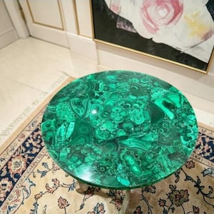 Puede incluir: Una mesa redonda con una vibrante tapa de malaquita verde. La superficie de la mesa presenta un patrón arremolinado de tonos verdes oscuros y claros. La mesa está sobre una alfombra estampada con una combinación de colores beige y azul marino.
