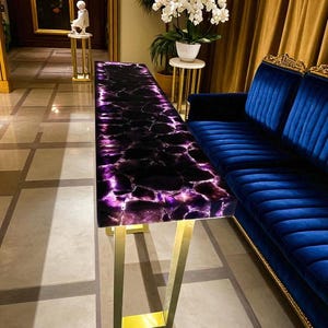 May include: A long, rectangular console table with a polished amethyst top and gold-colored metal legs. The table is positioned in a well-lit room with a blue velvet sofa and a painting on the wall. The amethyst top has a dark purple color.