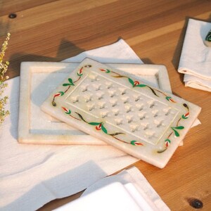 May include: A white marble soap dish with a rectangular shape and a decorative floral border in green, red, and black. The dish has a carved pattern of small, repeating shapes. A second, similar dish is visible.