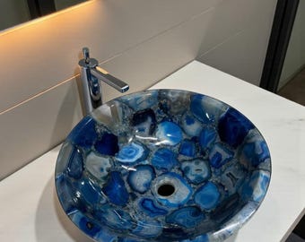 Pode incluir: Uma pia redonda de pedra de ágata azul e branca. Possui uma torneira cromada e está colocada sobre uma bancada branca. O interior da pia é um mosaico de pedras azuis e brancas, um elemento de casa de banho único.