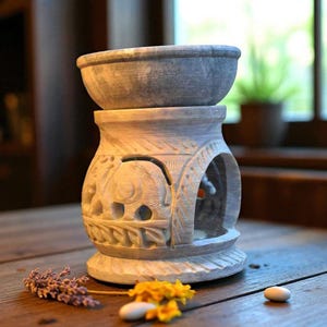 May include: A gray stone essential oil diffuser with an elephant carving. The diffuser has a bowl-shaped top and a carved base with arched openings. The base is decorated with leaf and floral patterns. The diffuser sits on a wooden surface.