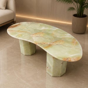 Op de afbeelding: Een salontafel gemaakt van lichtgroene onyxsteen, met een glad, onregelmatig ovaal blad met natuurlijke aders in tinten wit en amber, ondersteund door twee stevige, zeshoekige zuilen gemaakt van hetzelfde gepolijste steenmateriaal.