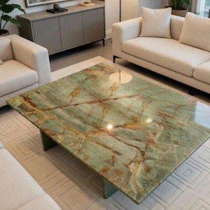May include: A square coffee table with a green marble top and a brown veined pattern. The table is set in a living room with cream-colored sofas and a patterned rug. The table's base is a dark green color.