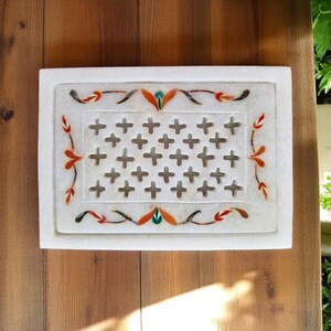 May include: A rectangular white marble decorative piece with a cross-shaped pattern in the center. The edges are adorned with colorful floral designs in orange, red, and green. The piece is set against a wooden surface.