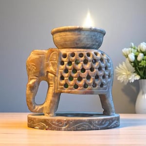 Könnte beinhalten: Ein geschnitzter Elefanten-Öl-Wärmer aus Stein mit einer brennenden Kerze. Der Elefant hat ein dekoratives Muster auf dem Rücken und eine Schale oben für Öl. Der Sockel ist ebenfalls mit einem dekorativen Muster versehen. Die Farbe ist hellbraun.
