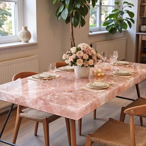 May include: A rectangular dining table with a polished pink quartz slab top and four wooden legs with black metal supports. The table is set with white plates, clear glasses, and napkins. A white vase with pink and white roses sits in the center.