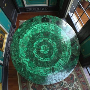 Può includere: Un grande tavolo rotondo con un piano in malachite verde vibrante. La superficie presenta motivi intricati e cerchi concentrici. Il tavolo è collocato in una stanza con un tappeto fantasia e una finestra.