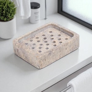 May include: A rectangular soap dish made of beige stone with multiple drainage holes. The soap dish is on a white countertop, with a small potted plant and a towel visible in the background. The design is simple and functional.