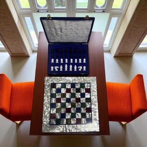 May include: A chess set with a wooden board and colorful squares, displayed on a dark wood table. The chess pieces are in a blue case. Two orange armchairs are on either side of the table.