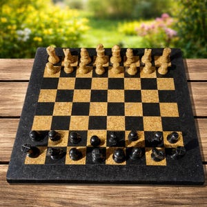 Puede incluir: Un juego de ajedrez con un tablero a cuadros negro y dorado y piezas de ajedrez negras y beige. El tablero está hecho de un material de piedra oscura, y las piezas están dispuestas para un juego. El fondo es una vista borrosa de un jardín.