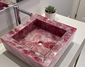 Può includere: Un lavabo rettangolare in pietra color rosa con un bacino quadrato. Il lavabo ha una finitura lucida e uno scarico centrale. Un rubinetto cromato è visibile, insieme a una piccola pianta succulenta in vaso.