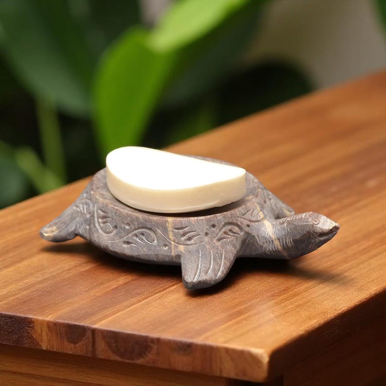 Puede incluir: Un jabonera gris en forma de tortuga de piedra con un dise&ntilde;o floral tallado, que sostiene una pastilla de jab&oacute;n blanca. La jabonera est&aacute; sobre una superficie de madera, con un fondo verde borroso.