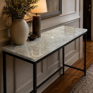 May include: A rectangular console table featuring a polished, light-colored stone top with a subtle crystalline pattern, supported by a slim, black metal frame. The table surface is smooth and reflective, showcasing natural mineral variations.