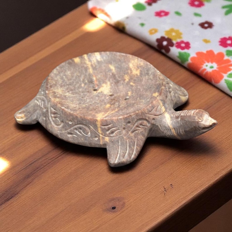 Puede incluir: Un jabonera con forma de tortuga de piedra gris y beige. El plato tiene un dise&ntilde;o tallado en los lados y est&aacute; sobre una superficie de madera. La cabeza de la tortuga apunta hacia la derecha.