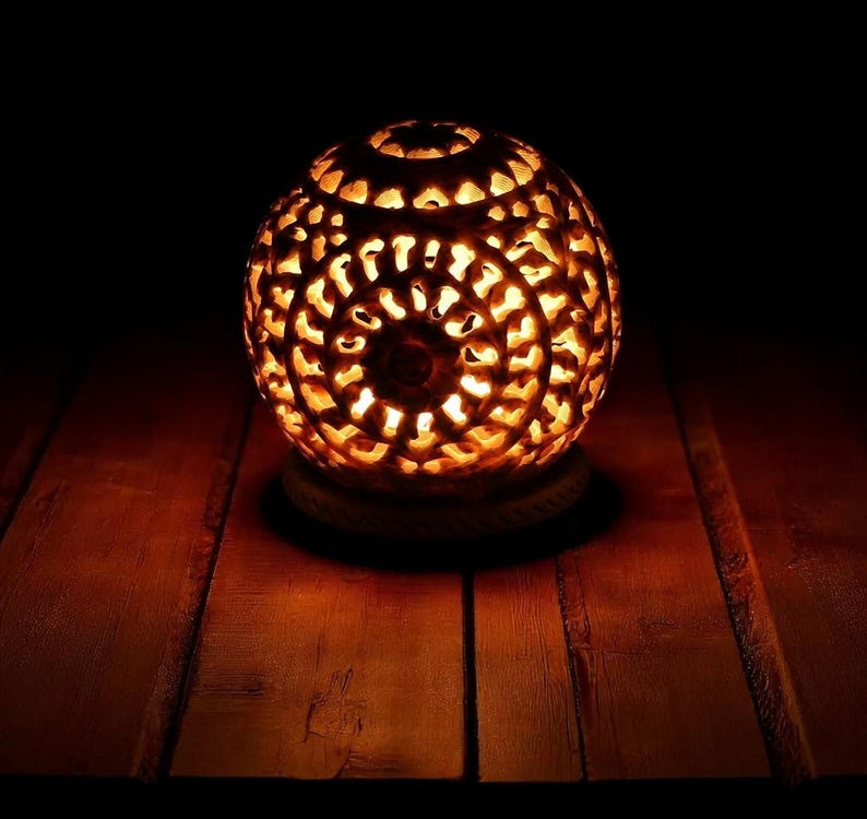 May include: A spherical, carved candle holder with intricate patterns, emitting a warm, orange glow. The decorative piece sits on a round, rope-textured base, creating a cozy ambiance. The background is dark, highlighting the illuminated design.