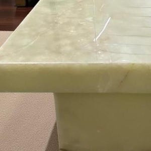 Puede incluir: Una mesa de centro cuadrada de &oacute;nix verde claro. La mesa tiene una base rectangular s&oacute;lida y una parte superior pulida con vetas naturales. La mesa est&aacute; sobre una alfombra de color neutro. Las dimensiones de la mesa no son visibles.