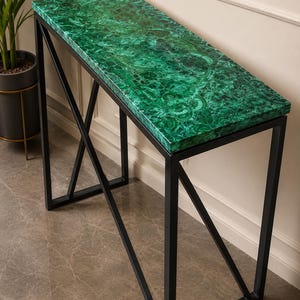 Puede incluir: Una mesa consola rectangular con un tablero de patrón de malaquita verde vibrante con texturas en espiral. La mesa está sostenida por un marco de metal negro construido con patas rectas y un diseño de refuerzo en cruz en los laterales.