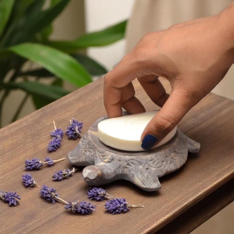 Puede incluir: Un jabonera gris en forma de tortuga de piedra sostiene una pastilla de jab&oacute;n blanca. La jabonera est&aacute; sobre una superficie de madera, con ramitas de lavanda morada esparcidas. La jabonera tiene detalles tallados.