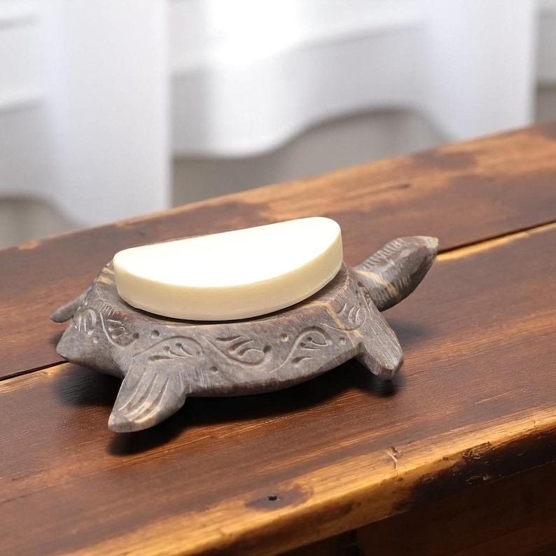 Puede incluir: Un jabonera de tortuga de piedra tallada con una pastilla de jab&oacute;n. La jabonera es gris y tiene un dise&ntilde;o detallado. El jab&oacute;n es de color crema claro. La jabonera est&aacute; sobre una superficie de madera.