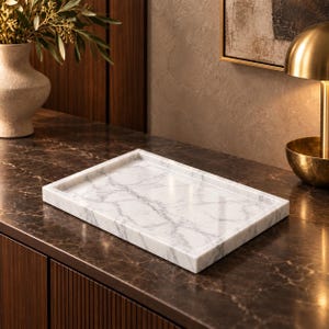 May include: A rectangular tray crafted from white marble with prominent grey veining. The tray features a smooth, polished surface and raised edges, providing a flat, stable base for organizing small items on a flat surface.