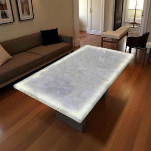 May include: A rectangular, white marble-topped coffee table with a glowing edge. The table is supported by a gray, rectangular base. The table is in a living room with a brown sofa and hardwood floors.