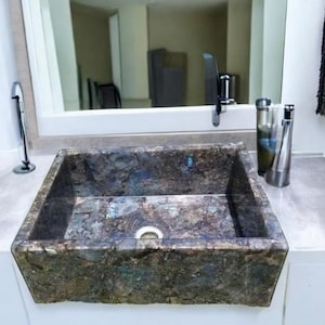May include: A square, stone bathroom sink with a textured, multi-colored surface. The sink is set into a white countertop, with a chrome faucet and a mirror in the background. A soap dispenser and a bottle are on the counter.