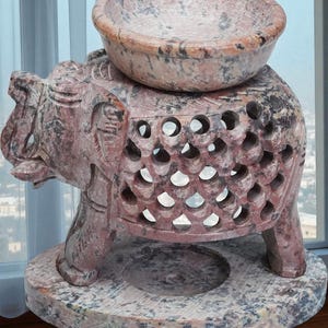 May include: A reddish-brown carved stone elephant sculpture with a bowl on its back. The elephant has intricate cut-out designs and stands on a round base. The sculpture's color is accented with dark grey and black.