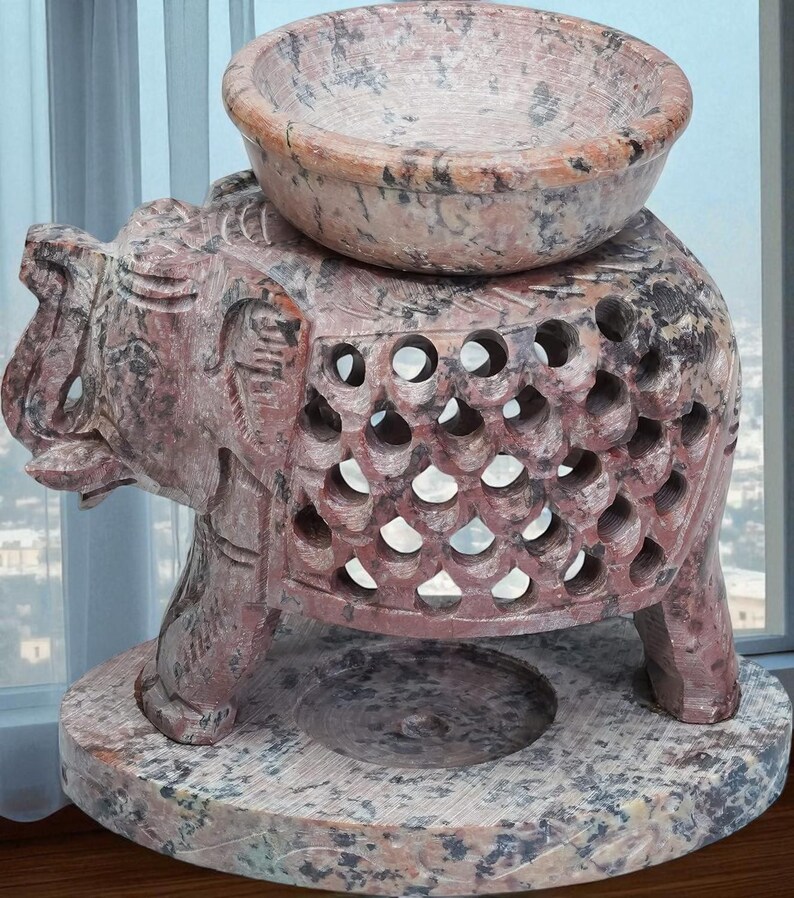 May include: A reddish-brown carved stone elephant sculpture with a bowl on its back. The elephant has intricate cut-out designs and stands on a round base. The sculpture's color is accented with dark grey and black.
