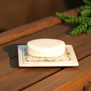 May include: A white oval bar of soap rests on a rectangular soap dish. The soap dish is white with a floral design in green and red. The soap and dish are on a wooden surface.