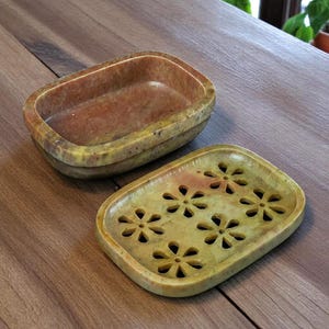 May include: Two-piece soap dish set. The base is a rectangular dish with a raised rim. The top piece is a matching dish with flower-shaped cutouts. The soap dishes are made of a mottled tan and brown stone.