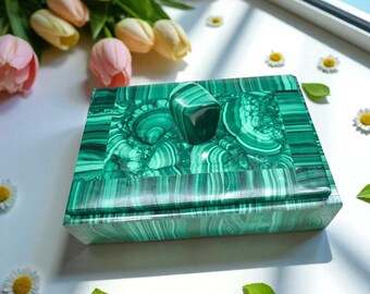 May include: A green malachite box with a matching stone, set against a backdrop of pink and yellow tulips and small white daisies. The box features a distinctive swirling pattern.