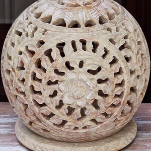 May include: A carved stone sphere with intricate floral and leaf patterns. The beige orb sits on a matching circular base. The detailed openwork design allows light to pass through, creating a decorative accent piece.