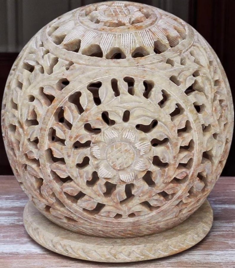 May include: A carved stone sphere with intricate floral and leaf patterns. The beige orb sits on a matching circular base. The detailed openwork design allows light to pass through, creating a decorative accent piece.
